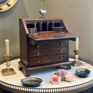 Può includere: Una piccola scrivania antica in legno con una superficie di scrittura incernierata e tre cassetti. Una piccola statuetta di uccello è appoggiata sulla scrivania. La scrivania è su un tavolo con piano in marmo, con candele e ciotole decorative. Rose rosa e viola sono anche sul tavolo.