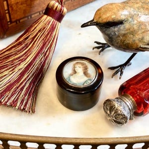 Puede incluir: Naturaleza muerta con objetos antiguos: un pájaro decorativo, una pequeña caja redonda con un retrato, una botella de vidrio roja y una borla. Los objetos están dispuestos sobre una superficie blanca, con una bandeja dorada.