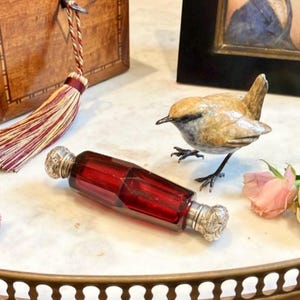 Puede incluir: Un frasco de perfume de vidrio rojo con tapones plateados ornamentados. Una pequeña y detallada figura de pájaro está cerca. Una borla decorativa y un marco de fotos también son visibles sobre una superficie de mármol.