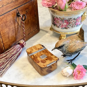 May include: A rectangular box with a mottled brown and tan tortoiseshell-like pattern, featuring silver-toned metal accents on the lid and a small central silver plaque, positioned near a bronze bird figurine and a floral porcelain container.