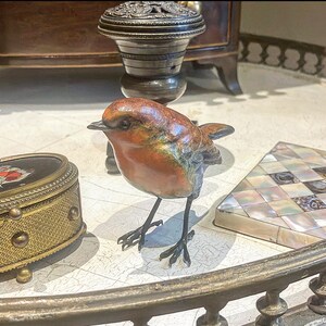 Puede incluir: Una peque&ntilde;a escultura de bronce de un petirrojo con el pecho rojo, posada sobre una mesa con una incrustaci&oacute;n decorativa. El p&aacute;jaro tiene una apariencia realista y detallada.