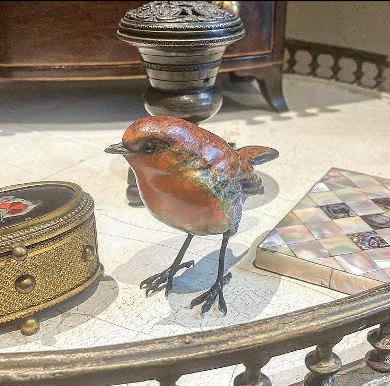Puede incluir: Una peque&ntilde;a escultura de bronce de un petirrojo con el pecho rojo, posada sobre una mesa con una incrustaci&oacute;n decorativa. El p&aacute;jaro tiene una apariencia realista y detallada.