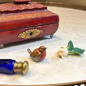 May include: A red wooden box with gold accents and a small porcelain bird figurine. The box is on a white marble surface with a blue glass bottle and a white rose.