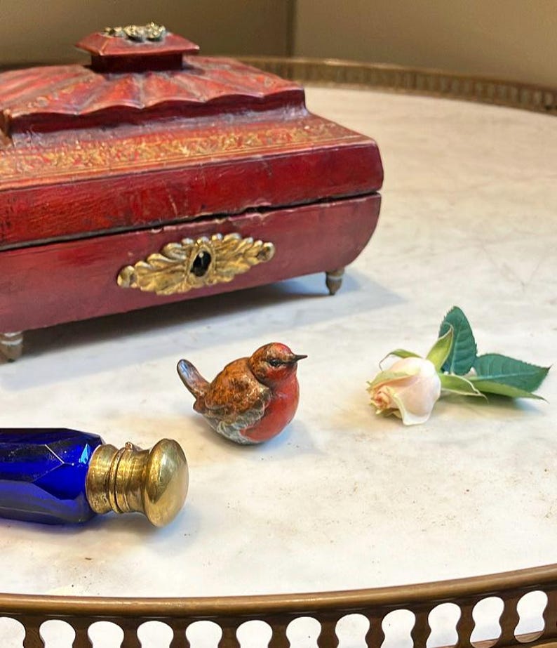 May include: A red wooden box with gold accents and a small porcelain bird figurine. The box is on a white marble surface with a blue glass bottle and a white rose.