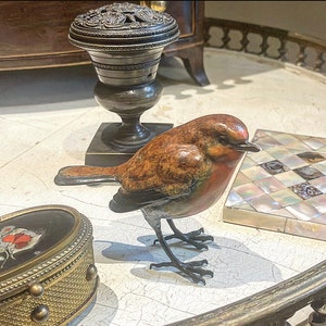 Puede incluir: Una escultura de bronce de un petirrojo con plumas marrones y naranjas. El p&aacute;jaro est&aacute; posado sobre una superficie blanca con una bandeja decorativa y un jarr&oacute;n de bronce en el fondo.