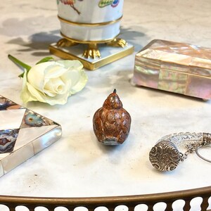 May include: A small, brown, ceramic bird figurine with a textured surface. The bird is sitting on a white marble surface.