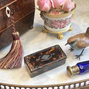 May include: A collection of antique items on a marble surface. Includes a tortoiseshell box, a small porcelain vase with pink roses, a bronze bird figurine, a blue glass bottle, and a decorative tassel.