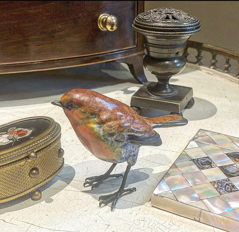 Puede incluir: Una peque&ntilde;a escultura de bronce detallada de un petirrojo con el pecho rojo y plumas marrones, negras y naranjas. El p&aacute;jaro est&aacute; posado sobre una superficie blanca.