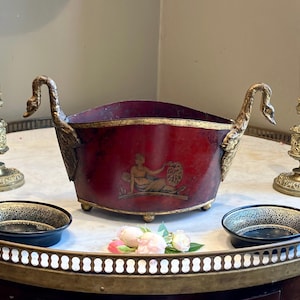 May include: A deep red oval metal bowl featuring gold-toned swan-shaped handles on each side and a gold-painted classical figure illustration on the front. The bowl has a gold-colored rim and rests on small feet, with a weathered finish throughout.