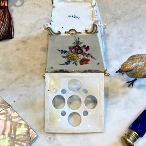 May include: A white porcelain flower frog with a square base featuring five circular openings of varying sizes. The base is decorated with a colorful floral bouquet and gold trim along the edges, resting on four small, ornate, gold-accented feet.