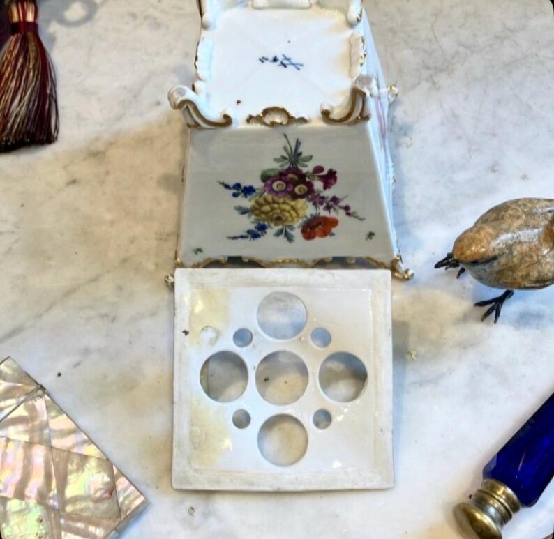 May include: A white porcelain flower frog with a square base featuring five circular openings of varying sizes. The base is decorated with a colorful floral bouquet and gold trim along the edges, resting on four small, ornate, gold-accented feet.