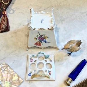 May include: A white porcelain flower frog with a square base, four feet, and floral motifs. The matching lid features five circular openings and two curved handles. Nearby are a small brown bird figurine, a blue glass vial, and a mother-of-pearl box.