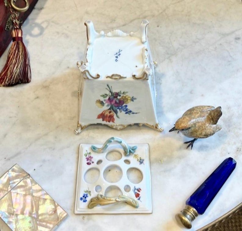 May include: A white porcelain flower frog with a square base, four feet, and floral motifs. The matching lid features five circular openings and two curved handles. Nearby are a small brown bird figurine, a blue glass vial, and a mother-of-pearl box.