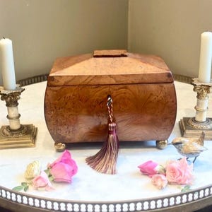 Puede incluir: Una caja de madera con un diseño curvo, con cerradura y borla decorativa. Dos velas blancas en candelabros ornamentados, rosas rosas y una pequeña figura de pájaro rodean la caja. La caja está sobre una mesa redonda.