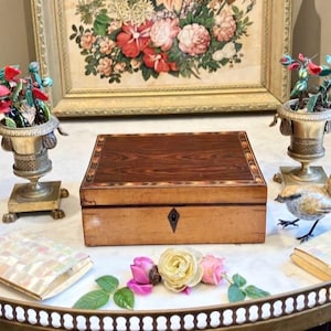 Peut inclure: Une boîte en bois rectangulaire avec un couvercle incrusté à motifs. Autour de la boîte, des objets décoratifs, dont de petits vases avec des compositions florales, un oiseau décoratif et un compact en nacre.