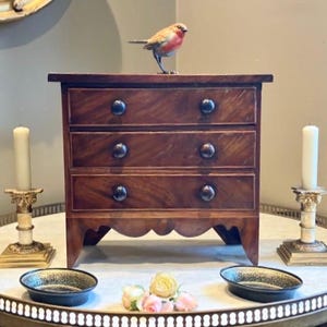 Può includere: Un piccolo comò in legno a tre cassetti con pomelli scuri. Una statuetta decorativa di uccello è appoggiata sopra il comò. Due candelabri con candele accese affiancano il comò. Il comò e l'arredamento sono su un tavolo rotondo.