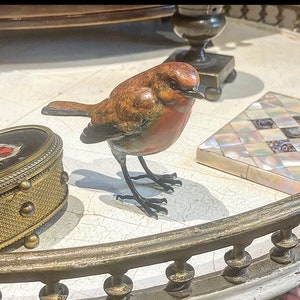 Puede incluir: Una peque&ntilde;a escultura de bronce detallada de un petirrojo con el pecho rojo y plumas marrones y naranjas. El p&aacute;jaro est&aacute; posado sobre una superficie blanca con un borde decorativo de metal dorado.