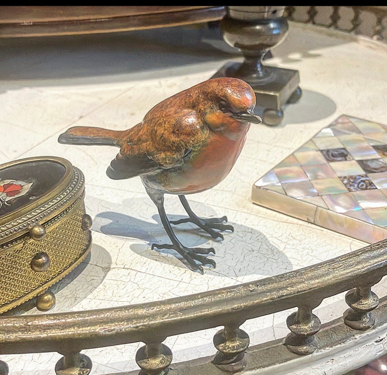 Puede incluir: Una peque&ntilde;a escultura de bronce detallada de un petirrojo con el pecho rojo y plumas marrones y naranjas. El p&aacute;jaro est&aacute; posado sobre una superficie blanca con un borde decorativo de metal dorado.