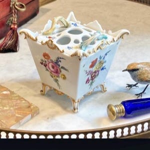 May include: A square white porcelain flower frog with a flared rim, gold-painted edges, and four feet. The sides feature hand-painted floral bouquets in pink, orange, yellow, and blue. The top has multiple circular openings for stems and two small handles.