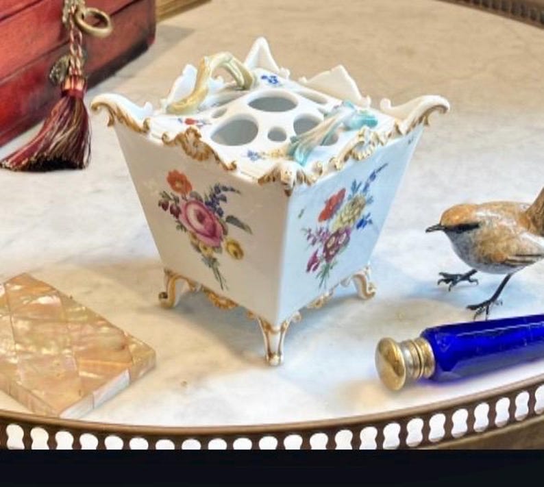 May include: A square white porcelain flower frog with a flared rim, gold-painted edges, and four feet. The sides feature hand-painted floral bouquets in pink, orange, yellow, and blue. The top has multiple circular openings for stems and two small handles.