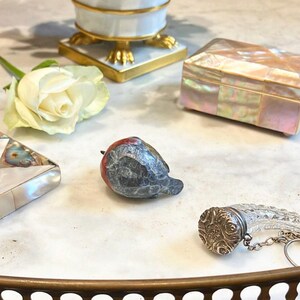 May include: A small, carved, gray and red bird figurine with a white rose and a mother of pearl box in the background.
