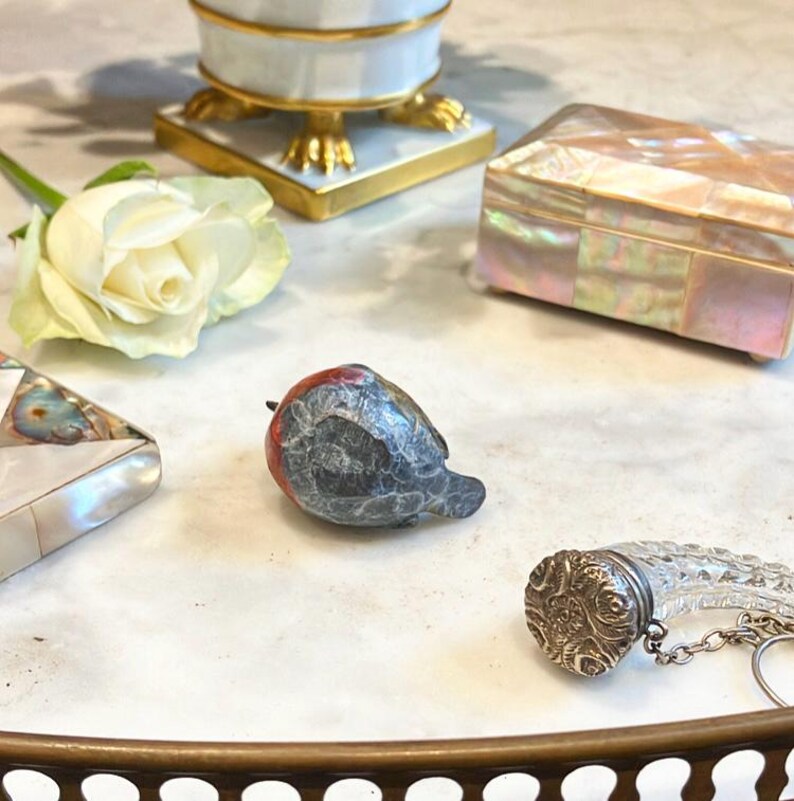 May include: A small, carved, gray and red bird figurine with a white rose and a mother of pearl box in the background.