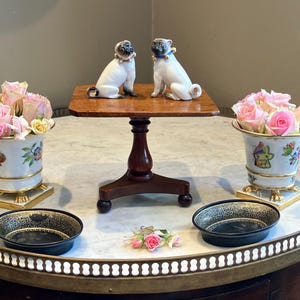 Può includere: Due statuette di carlino in porcellana con collare poggiano su un piedistallo in legno. Due vasi in ceramica bianca con motivi floreali e farfalle contengono rose rosa. Due ciotole nere poco profonde con motivi in filigrana dorata sul tavolo.