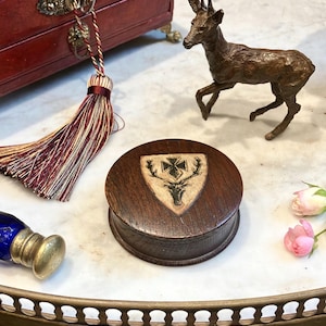 May include: A round dark brown wooden box featuring a light-colored shield-shaped inlay on the lid. The inlay displays a black graphic of a stag head topped with a cross. The box has a smooth, polished finish and a circular shape.