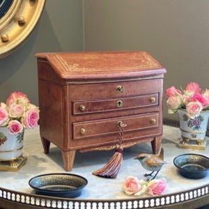 Può includere: Un piccolo forziere antico in legno con tre cassetti, un piano inclinato e ferramenta in ottone. Il forziere è marrone con accenti dorati e poggia su un tavolo con piano in marmo. Rose rosa in vasi decorativi e una piccola statuina di uccello sono anche sul tavolo.