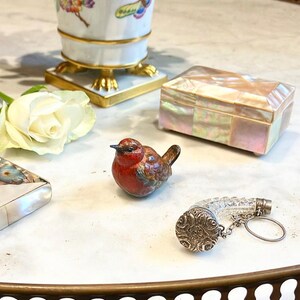May include: A small, red and brown bird figurine sits on a white surface.  A clear glass bottle stopper with a silver cap and chain is next to the bird.  A white rose and a small, iridescent box are also in the image.