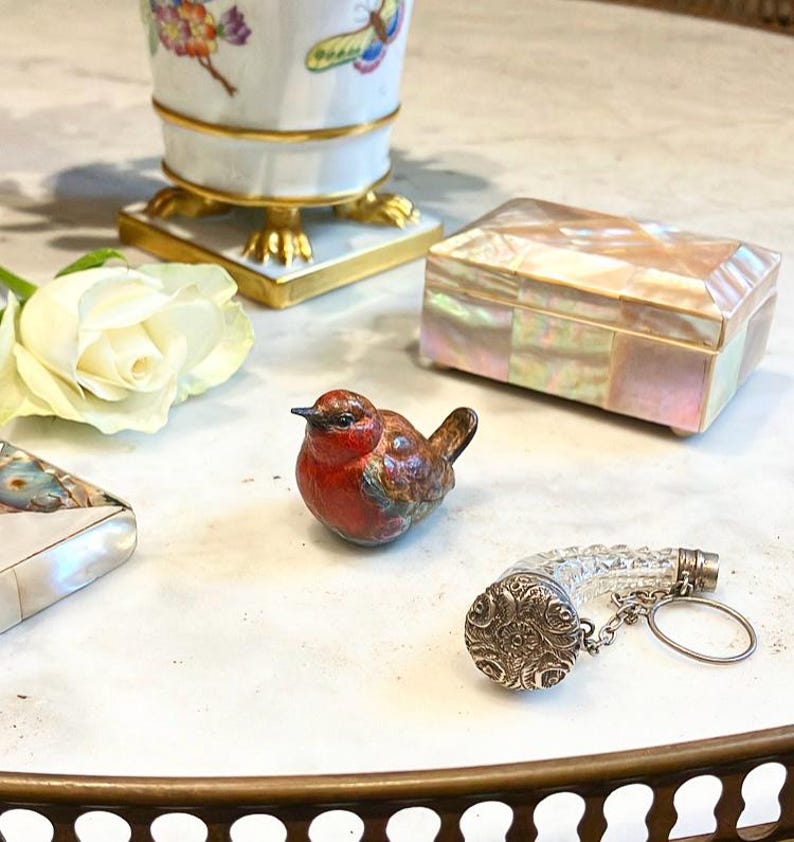 May include: A small, red and brown bird figurine sits on a white surface.  A clear glass bottle stopper with a silver cap and chain is next to the bird.  A white rose and a small, iridescent box are also in the image.