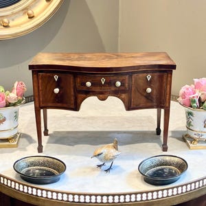 Può includere: Un piccolo mobile antico in legno con fronte curvo e tre cassetti. Il mobile è in legno scuro con dettagli intarsiati e poggia su quattro gambe sottili. Vasi di fiori decorativi fiancheggiano il mobile e una piccola statuina di uccello è di fronte.