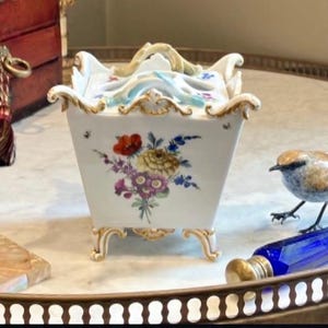 May include: A square white porcelain container with a lid, featuring hand-painted floral bouquets in red, yellow, and purple. The piece has ornate gold-trimmed edges, four decorative feet, and a sculpted handle on top with light blue accents.