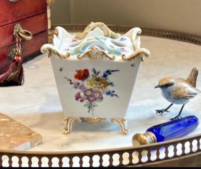May include: A square white porcelain container with a lid, featuring hand-painted floral bouquets in red, yellow, and purple. The piece has ornate gold-trimmed edges, four decorative feet, and a sculpted handle on top with light blue accents.