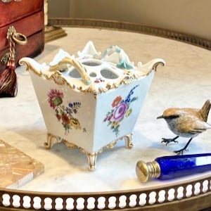 May include: A white porcelain flower frog with a square, tapered body, four ornate feet, and gold-trimmed edges. The top features multiple circular openings and delicate floral motifs in pink, orange, blue, and green painted on the sides.