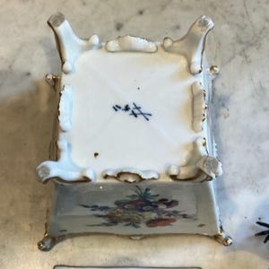 May include: A square white porcelain container with four ornate feet and gold-painted edges. The base features a blue crossed-swords maker's mark. The side displays a colorful floral arrangement with yellow, purple, and red blossoms on a white surface.