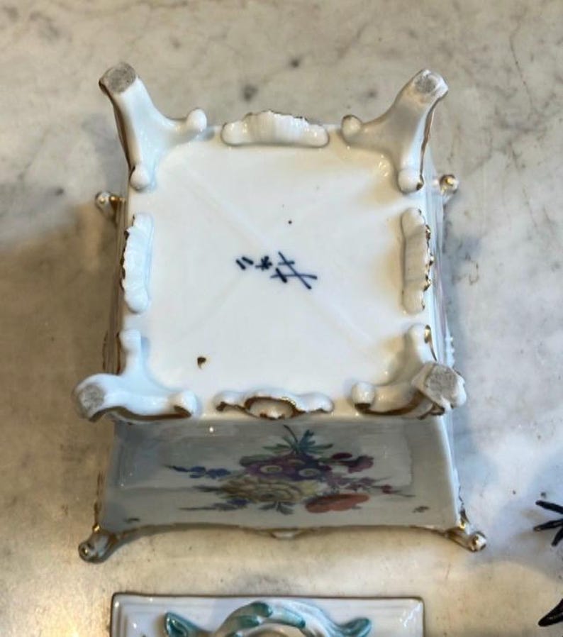 May include: A square white porcelain container with four ornate feet and gold-painted edges. The base features a blue crossed-swords maker's mark. The side displays a colorful floral arrangement with yellow, purple, and red blossoms on a white surface.