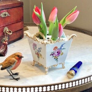 May include: A white ceramic flower frog vase with gold-trimmed edges and feet, featuring hand-painted floral motifs on the sides. The vase contains several pink tulips with green stems and leaves, arranged in the top openings of the vessel.