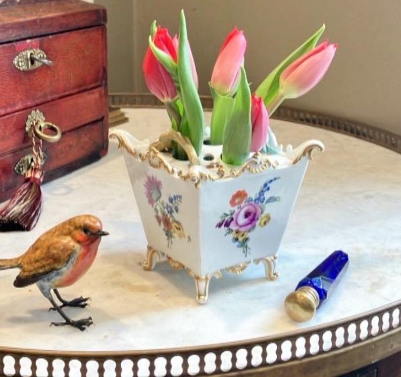 May include: A white ceramic flower frog vase with gold-trimmed edges and feet, featuring hand-painted floral motifs on the sides. The vase contains several pink tulips with green stems and leaves, arranged in the top openings of the vessel.