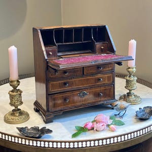 Può includere: Uno scrittoio da tavolo in legno con piano a ribalta con interno rosso, due piccoli cassetti superiori e tre cassetti inferiori più grandi con pomelli in metallo e una serratura centrale, affiancato da due candelabri in ottone con candele rosa.