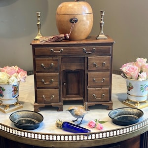 Può includere: Un organizer da scrivania in legno con sette cassetti e un'anta centrale, sormontato da un grande contenitore a forma di mela, due candelabri in ottone, due vasi in porcellana floreale, due ciotole nere, una statuetta di uccello e una bottiglia blu.