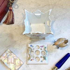 May include: A white porcelain square flower frog vase with gold trim and floral motifs, accompanied by a matching perforated lid with five circular openings, two decorative side handles, and four small feet on the base of the vessel.