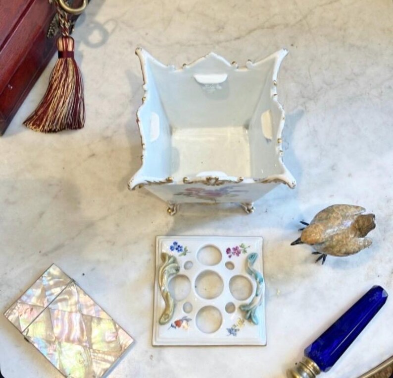 May include: A white porcelain square flower frog vase with gold trim and floral motifs, accompanied by a matching perforated lid with five circular openings, two decorative side handles, and four small feet on the base of the vessel.