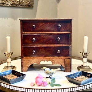 Può includere: Un piccolo comò antico in legno con tre cassetti. Il comò è realizzato in legno scuro con un ricco motivo a venature. Due candele bianche in portacandele ornati affiancano il comò. Vassoi decorativi e una statuina di uccello sono sul tavolo.