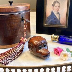 May include: A carved wooden monkey head figurine with dark inset eyes, positioned next to a cylindrical wooden box with a key and a decorative tassel, a framed portrait of a person in a blue coat, a cobalt blue glass bottle, and two small flowers.