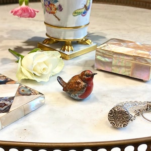 May include: A small, hand-painted ceramic bird figurine with brown, red, and white details. The bird is sitting on a white marble surface with a white rose, a porcelain vase with a butterfly design, and a mother-of-pearl box.