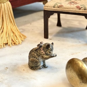 Può includere: Una piccola statuetta in metallo usurato raffigurante un gatto seduto che si pulisce la zampa. Il gatto presenta una finitura testurizzata nei toni del grigio e del marrone con segni visibili di usura, orecchie a punta e coda ripiegata.
