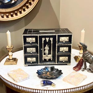 May include: A black and ivory wooden box with six drawers and a central door. The box is decorated with intricate carvings and a scene of a man in a hat and coat. The box is sitting on a white table with other decorative items.