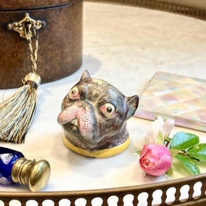May include: A ceramic pug dog figurine with a black and gray coat, pink nose, and yellow collar. A small pink rose and a blue glass bottle with a gold cap are also present. The items are arranged on a white surface.