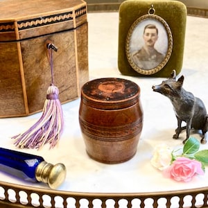 Puede incluir: Varios objetos antiguos sobre una superficie blanca. Incluye una caja de madera, un retrato en un marco de terciopelo verde, una figura de zorro de bronce, un pequeño recipiente de madera en forma de barril y una botella de vidrio azul.